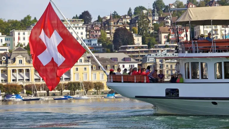 Bateau de passagers avec un drapeau suisse, devant des maisons au bord de l’eau.